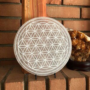 May include: A white selenite crystal plate with a carved flower of life design. The plate is on a stand and is in front of a brick wall.