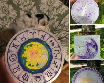 May include: Four round, white, decorative plates with purple and gold accents. One plate features a sun and moon design with the zodiac symbols around the edge. Another plate features a purple cat design. The third plate features a purple wolf design. The fourth plate features a purple moon design with a circle of purple dots around the edge.