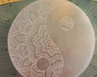 May include: A white selenite disc with a yin yang design etched into the surface. The design features intricate white mandala patterns on one side and a simple mandala on the other side.