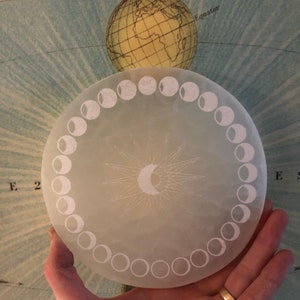 May include: A white circular moon phase tracker with a detailed sunburst design in the center. The tracker has 12 moon phases etched around the edge.