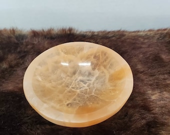 May include: A small, round, orange-colored bowl made of selenite crystal. The bowl has a smooth, polished surface and a slightly rippled texture.