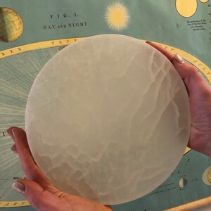May include: A white, frosted glass moon model with a textured surface. The model is being held by a person's hands. The model is in front of a vintage educational chart about astronomy.