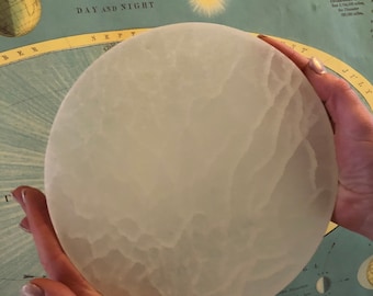 May include: A white, frosted glass moon model with a textured surface. The model is being held by a person's hands. The model is in front of a vintage astronomy chart with the text "ASTRONOMY" and "FIG. 1. DAY AND NIGHT".