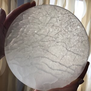 May include: A large, round, white selenite crystal plate with a natural, textured surface. The crystal is translucent and has a slight shimmer.