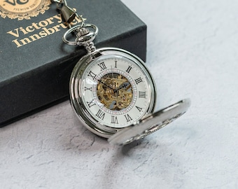 May include: A silver pocket watch with a white face and Roman numeral hour markers. The watch is open and the intricate inner workings are visible. The watch is on a chain and in a black box with the text "V&A Victory Innsbruck" on the top.