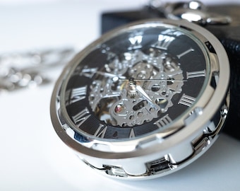 Op de afbeelding: Een zilveren zakhorloge met een zwarte wijzerplaat en Romeinse cijfers voor de uren. Het horloge heeft een skeletontwerp, waardoor de ingewikkelde interne mechanismen van het uurwerk zichtbaar zijn.