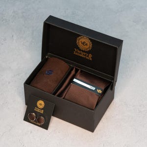 Könnte beinhalten: Schwarze Geschenkbox mit brauner Krawatte, Einstecktuch und Krawattenklammer. Die Box enthält auch ein Paar Manschettenknöpfe auf einer kleinen Karte. Der Markenname "Victory & Innsbruck" ist auf der Box und Karte aufgedruckt.