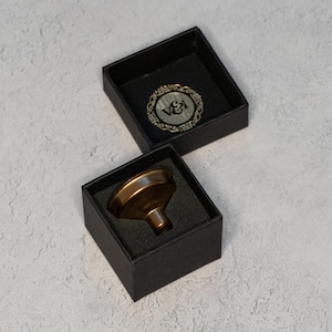 Peut inclure: Un entonnoir en métal doré dans une boîte noire avec un insert en mousse noire. La boîte porte un logo sur le dessus qui dit "V&I".