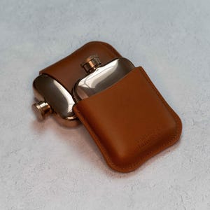 May include: Two stainless steel hip flasks with gold-coloured caps and brown leather cases. One flask is partially inside its case, and the other is next to it. The case has the text "Victory of Sunshine" embossed on it.