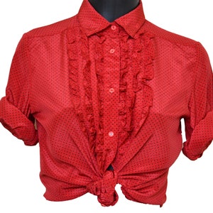 Könnte beinhalten: Eine rote Blusen mit weißen Punkten, einem Rüschen-Vorderteil und langen Ärmeln. Die Bluse ist vorne gebunden.