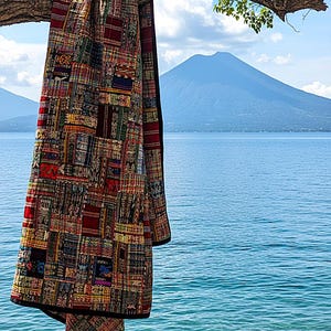 May include: A colorful patchwork quilt hangs from a tree branch against a backdrop of a lake and mountain. The quilt features a variety of patterned fabric squares in red, blue, yellow, and green. The quilt's edges are trimmed with a black border.