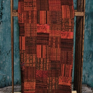 May include: A large, rectangular tapestry woven with a patchwork of colorful geometric patterns in shades of red, brown, and black. The tapestry is hanging on a wooden rack.