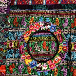 May include: A colorful textile with intricate embroidery featuring a circular design with flowers and figures. The textile is made up of various geometric patterns and vibrant colors, including red, green, blue, yellow, and purple.