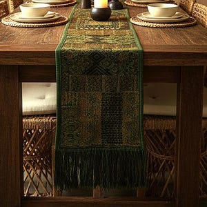 May include: A rectangular table runner featuring a dark green and gold woven pattern with geometric motifs and a fringed edge. The runner is centered on a wooden dining table set with woven placemats, ceramic bowls, and a central candle holder.