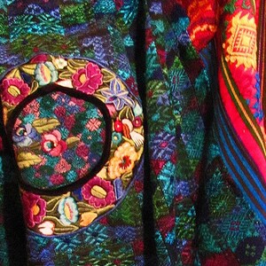 May include: A colorful textile with a large embroidered circle featuring flowers and leaves in shades of pink, blue, green, and yellow. The textile is woven with a geometric pattern in shades of blue, green, and red.