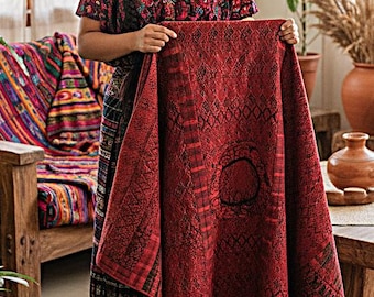 Op de afbeelding: Een rechthoekig textiel met een dieprode basiskleur, ingewikkelde zwarte geometrische patronen en een centraal cirkelvormig motief. De stof heeft een geweven textuur met verticaal gestreepte randen en een consistent rood en zwart kleurenpalet.