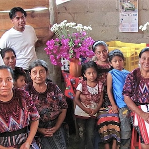 May include: A group of people, including children, are gathered together in a room. They are all wearing traditional clothing, and some are holding flowers. The room is decorated with flowers and plants.