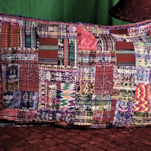 May include: A rectangular patchwork pillow with a variety of colorful fabric squares. The pillow features a mix of red, purple, and blue hues, with intricate patterns and textures. The pillow is resting on a dark brown surface.