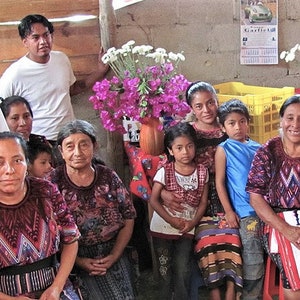 May include: A group of people, including children, are gathered together in a room. They are all wearing traditional clothing, and some are holding flowers. The room is decorated with flowers and plants.