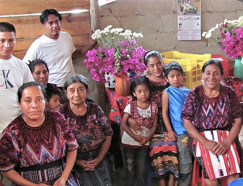 May include: A group of people, including children, are gathered together in a room. They are all wearing traditional clothing, and some are holding flowers. The room is decorated with flowers and plants.