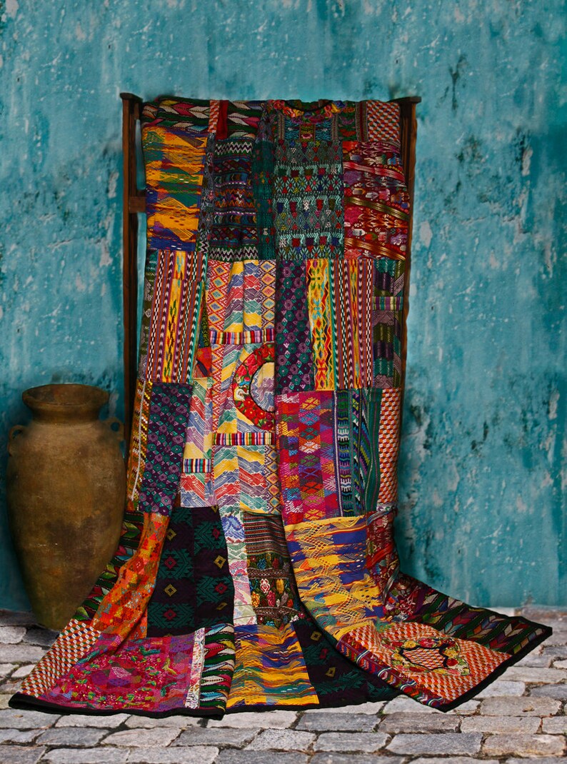 May include: A colorful patchwork quilt with a variety of geometric patterns and designs in shades of red, blue, yellow, green, and black. The quilt is draped over a wooden frame and is hanging against a turquoise wall.