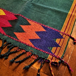 May include: A green woven textile with a colorful geometric design. The design features zig-zag patterns in red, orange, yellow, and purple. The edges are finished with a woven border and knotted fringe.