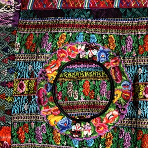 May include: A colorful textile with intricate embroidery featuring a circular design with flowers and figures. The textile is made up of various geometric patterns and vibrant colors, including red, green, blue, yellow, and purple.