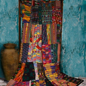 May include: A colorful patchwork quilt with a variety of geometric patterns and designs in shades of red, blue, yellow, green, and black. The quilt is draped over a wooden frame and is hanging against a turquoise wall.