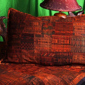 May include: A rectangular pillow with a patchwork design in shades of orange, brown, and black. The pillow has a textured appearance and is placed on a matching patterned bedspread. The overall theme is rustic and decorative.