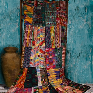 May include: A colorful patchwork quilt with a variety of geometric patterns and designs in shades of red, blue, yellow, green, and black. The quilt is draped over a wooden frame and is hanging against a turquoise wall.