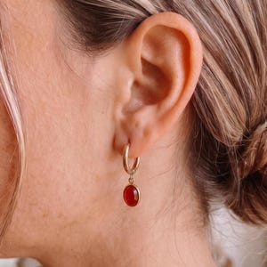 Könnte beinhalten: Ein goldener Creolen-Ohrring mit einem kleinen, glatten, ovalen roten Edelsteinanhänger, der an der Unterseite hängt. Der Ohrring ist am Ohrläppchen befestigt und zeichnet sich durch ein poliertes Metallfinish und einen leuchtend roten Cabochon-Stein aus.