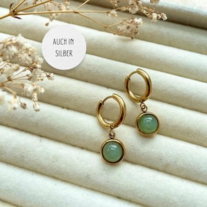 May include: A pair of gold hoop earrings with a green stone pendant. The earrings are displayed on a white background with the text "AUCH IN SILBER" on a white circle.