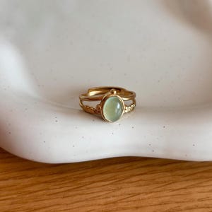 Op de afbeelding: Een goudkleurige ring met een gladde, ovaalvormige lichtgroene cabochonsteen in het midden. De ringband heeft een gespleten ontwerp met een getextureerd, touwachtig patroon aan de zijkanten en een verstelbare opening aan de achterkant.