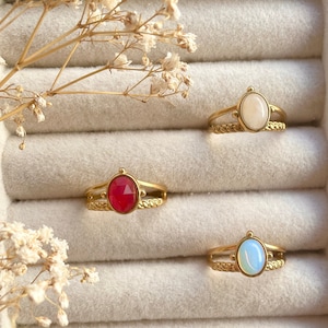 May include: Three gold rings with oval-shaped gemstones. The rings are displayed in a white velvet ring box. The gemstones are white, red, and blue.