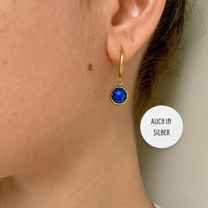 Op de afbeelding: Gouden creolen met een hangende blauwe edelsteen. De tekst "AUCH IN SILBER" is zichtbaar in de afbeelding.