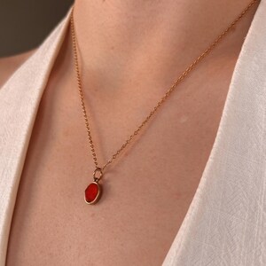 Può includere: Una collana a catena in metallo color oro con un piccolo ciondolo di forma ovale caratterizzato da una pietra centrale rossa liscia e vivace. Le maglie della catena sono uniformi e il ciondolo è fissato alla catena con un piccolo anello.