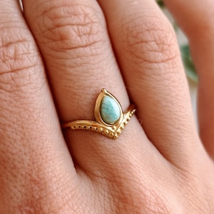Könnte beinhalten: Ein goldfarbener Ring mit einem tropfenförmigen, hellblauen Edelstein. Der Ring hat ein Chevron-Design mit kleinen, dekorativen Perlen entlang des Bandes. Der Ring befindet sich an einem Finger.