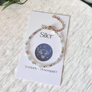 Op de afbeelding: Een delicate armband met kleine, lichtblauwe en heldere kralen afgewisseld met goudkleurige vierkante kralen. De armband wordt getoond op een witte kaart met het woord "Stier" en een Stier-dierenriemteken. De kaart bevat ook de tekst "Aventurin + Rosenquarz".