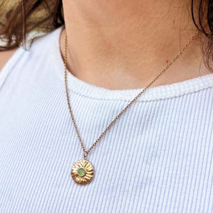 May include: A gold-toned metal necklace featuring a thin chain and a circular pendant with a textured, sunburst-like surface. The center of the pendant is set with a small, round, light green gemstone. The necklace rests against a white ribbed top.
