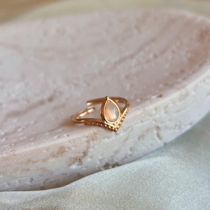 Könnte beinhalten: Ein goldfarbener Ring mit einem tropfenförmigen, irisierenden Stein. Der Ring hat ein V-förmiges Design mit kleinen Perlenakzenten. Der Ring wird auf einer weißen, ovalen Schale präsentiert.