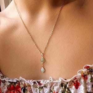 Op de afbeelding: Een gouden ketting met twee traanvormige stenen. De bovenste steen is lichtgroen en de onderste steen is lichtblauw.