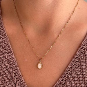 Op de afbeelding: Een goudkleurige ketting met een kleine, ovale hanger met een gladde, melkwitte cabochonsteen in een gepolijste goudkleurige metalen zetting. De fijne schakelketting rust op de hals en toont een eenvoudig en verfijnd ontwerp.