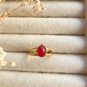 Könnte beinhalten: Ein goldfarbener Ring mit einem zentralen, facettierten roten Stein in Ovalform. Die Ringschiene besteht aus zwei Teilen: einem glatten oberen Draht und einem unteren Band mit einer texturierten, gedrehten Kordelstruktur, die an der Fassung zusammenlaufen.