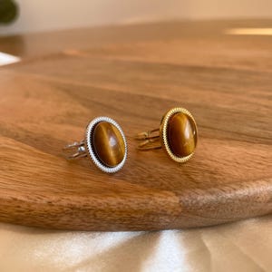 Könnte beinhalten: Zwei ovale Ringe mit einem braunen Tigerauge-Stein, der in einer goldenen und silbernen Fassung sitzt. Die Ringe liegen auf einer Holzoberfläche.