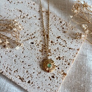 May include: A gold-toned necklace featuring a thin chain and a circular pendant with a textured, sunburst-like surface. The center of the pendant is set with a small, smooth, round light green stone. The metal has a polished, reflective finish.