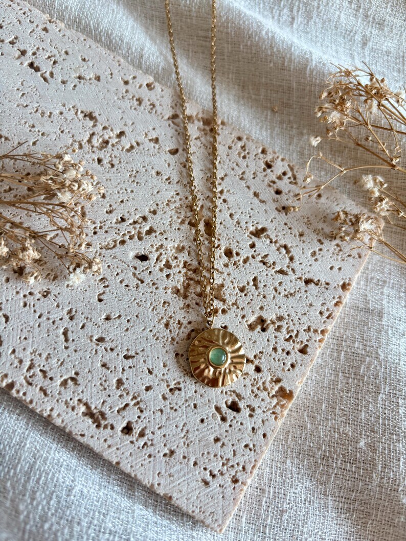 May include: A gold-toned necklace featuring a thin chain and a circular pendant with a textured, sunburst-like surface. The center of the pendant is set with a small, smooth, round light green stone. The metal has a polished, reflective finish.