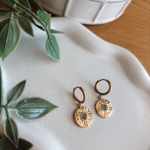 May include: A pair of gold-toned hoop earrings featuring circular, textured medallions. Each medallion has a sunburst-like pattern radiating from a small, round, light green stone set in the center. The earrings are designed with a hinged closure.