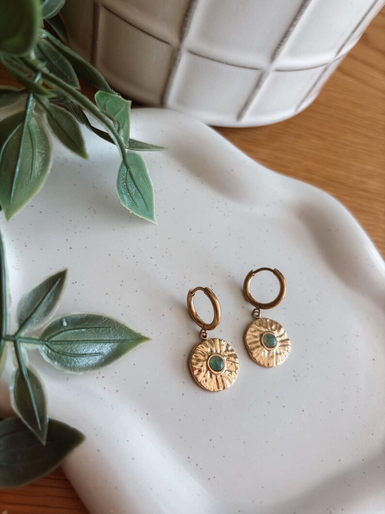 May include: A pair of gold-toned hoop earrings featuring circular, textured medallions. Each medallion has a sunburst-like pattern radiating from a small, round, light green stone set in the center. The earrings are designed with a hinged closure.