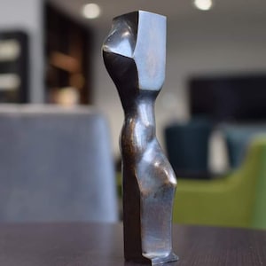 May include: Bronze abstract sculpture of a human form. The sculpture has a rectangular head and a slender, curved body. The bronze has a dark, aged patina, and the sculpture is displayed on a dark brown table.
