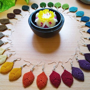 Puede incluir: Un conjunto de 15 pares de pendientes de macramé hechos a mano en un arcoíris de colores, incluyendo marrón, beige, amarillo, naranja, rojo, morado, azul y verde. Los pendientes tienen forma de hojas y se muestran sobre una superficie de madera.